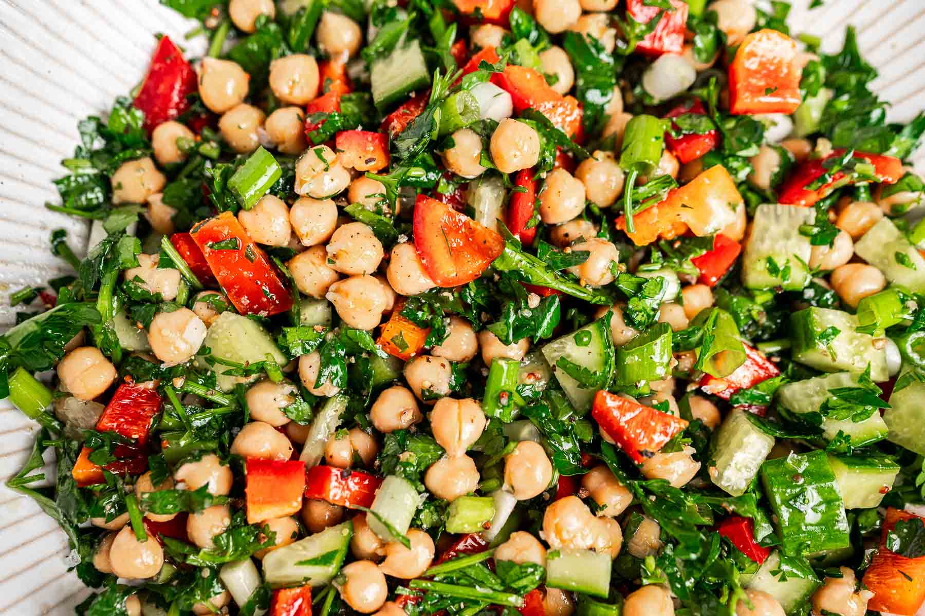 A bowl of chopped chickpea salad featuring red bell peppers, cucumbers, parsley, and fresh herbs.