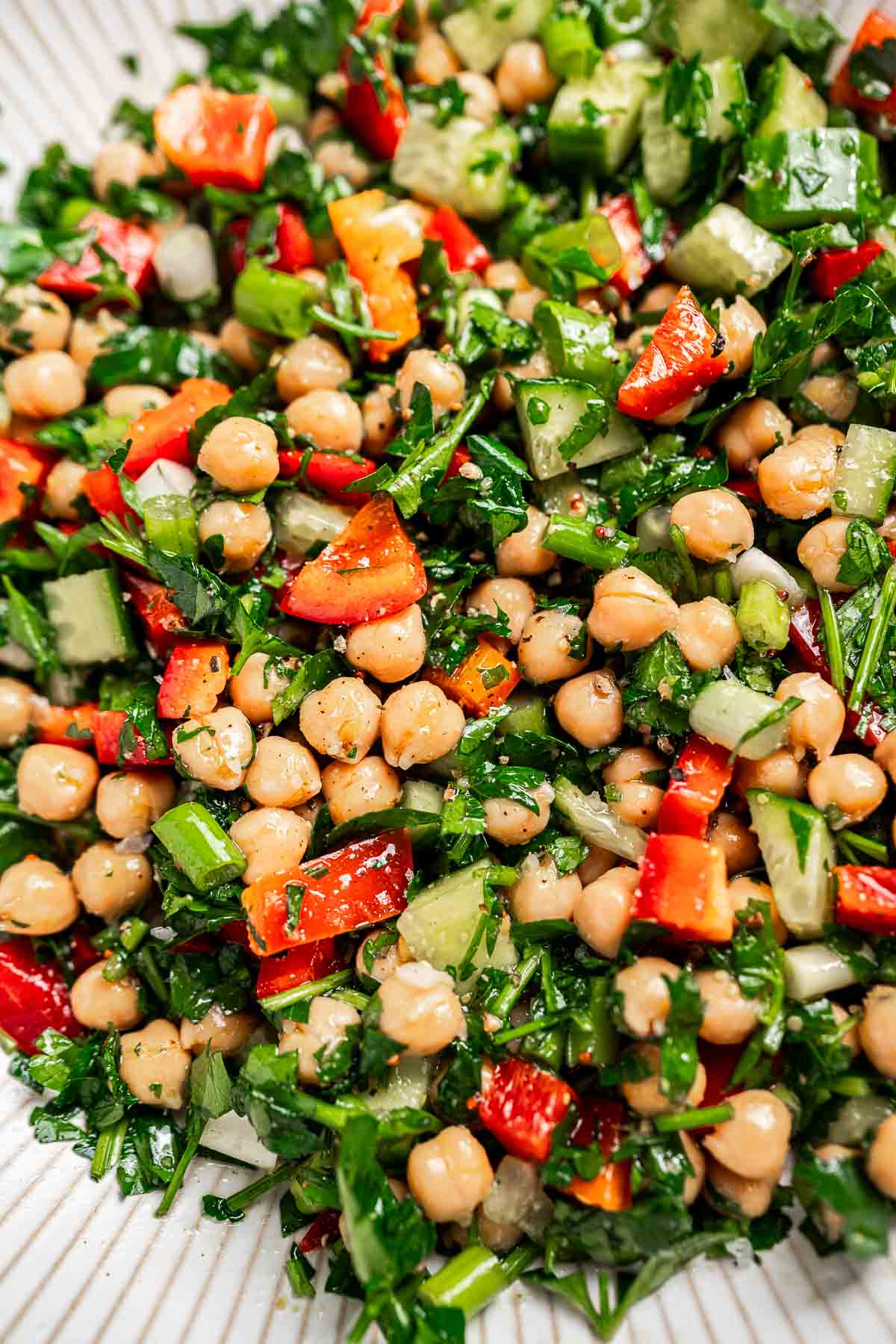 A vibrant chopped chickpea salad featuring a medley of colorful red and green peppers, crisp cucumbers, fresh parsley, and fragrant herbs, all beautifully presented on a pristine white plate.