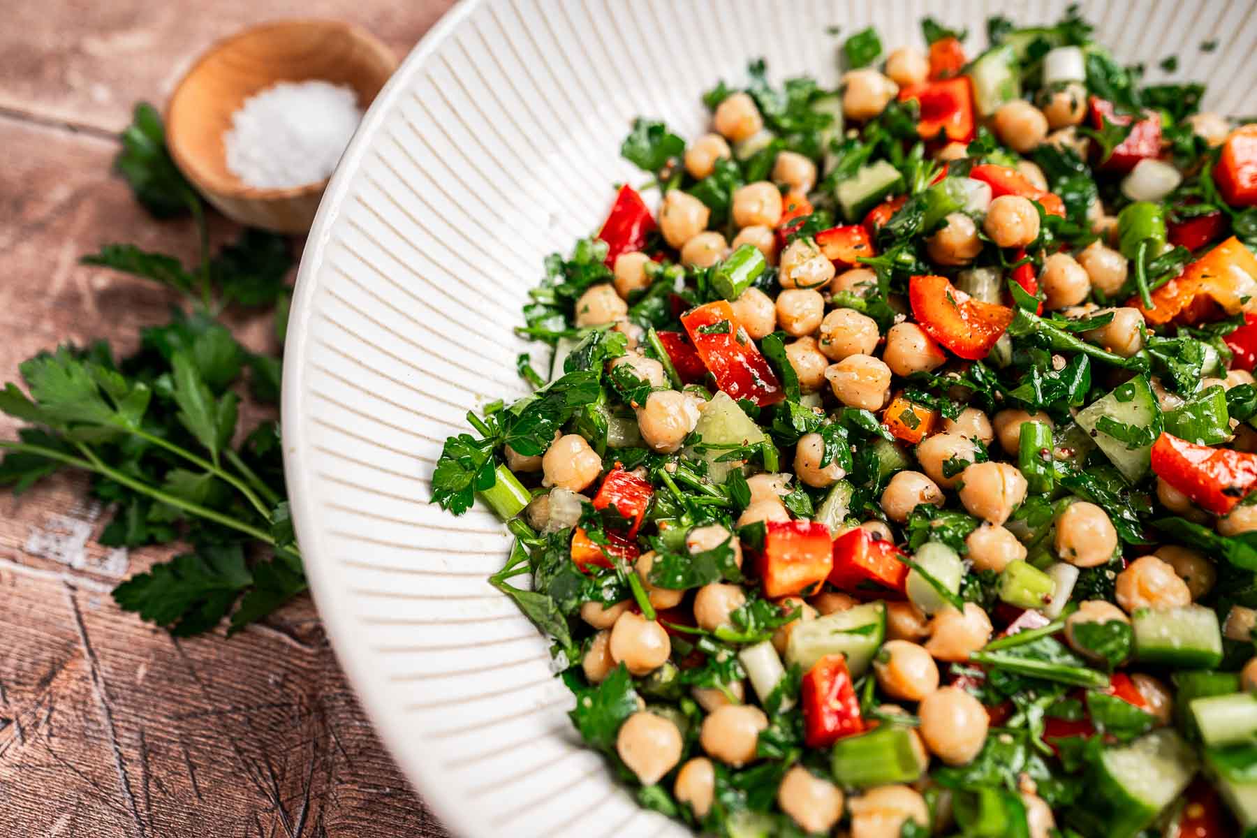 A bowl of chopped chickpea salad with fresh tomatoes, crisp cucumbers, and vibrant parsley sits on a wooden surface, with garnish nearby.