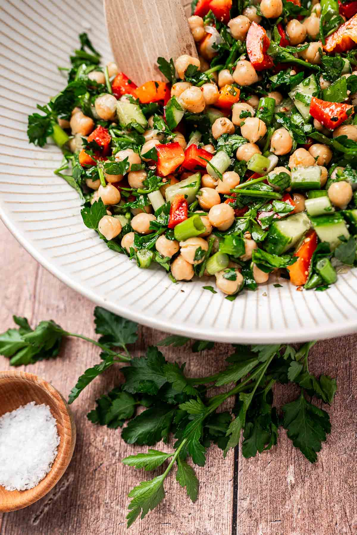 A bowl of chopped chickpea salad with cucumber, red bell pepper, and parsley sits on a wooden table beside a small bowl of salt and a sprig of fresh parsley.