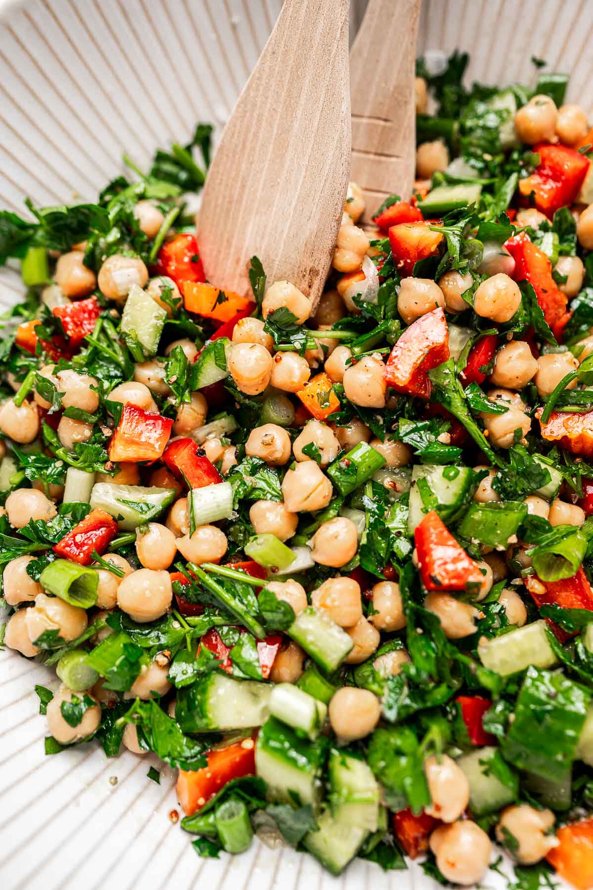 A chopped chickpea salad featuring red peppers, cucumber, and parsley tossed in a white bowl with wooden utensils.