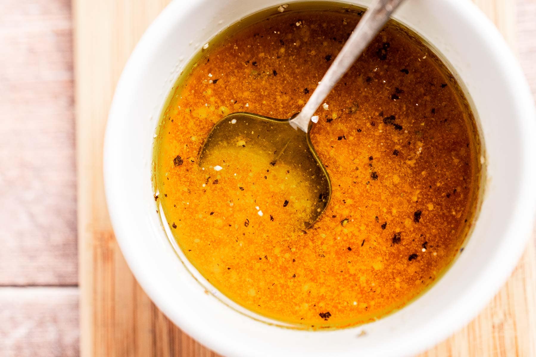 A bowl of golden orange dressing with black pepper flakes, perfect for drizzling over a refreshing chopped chickpea salad, with a spoon resting inside, placed on a wooden surface.