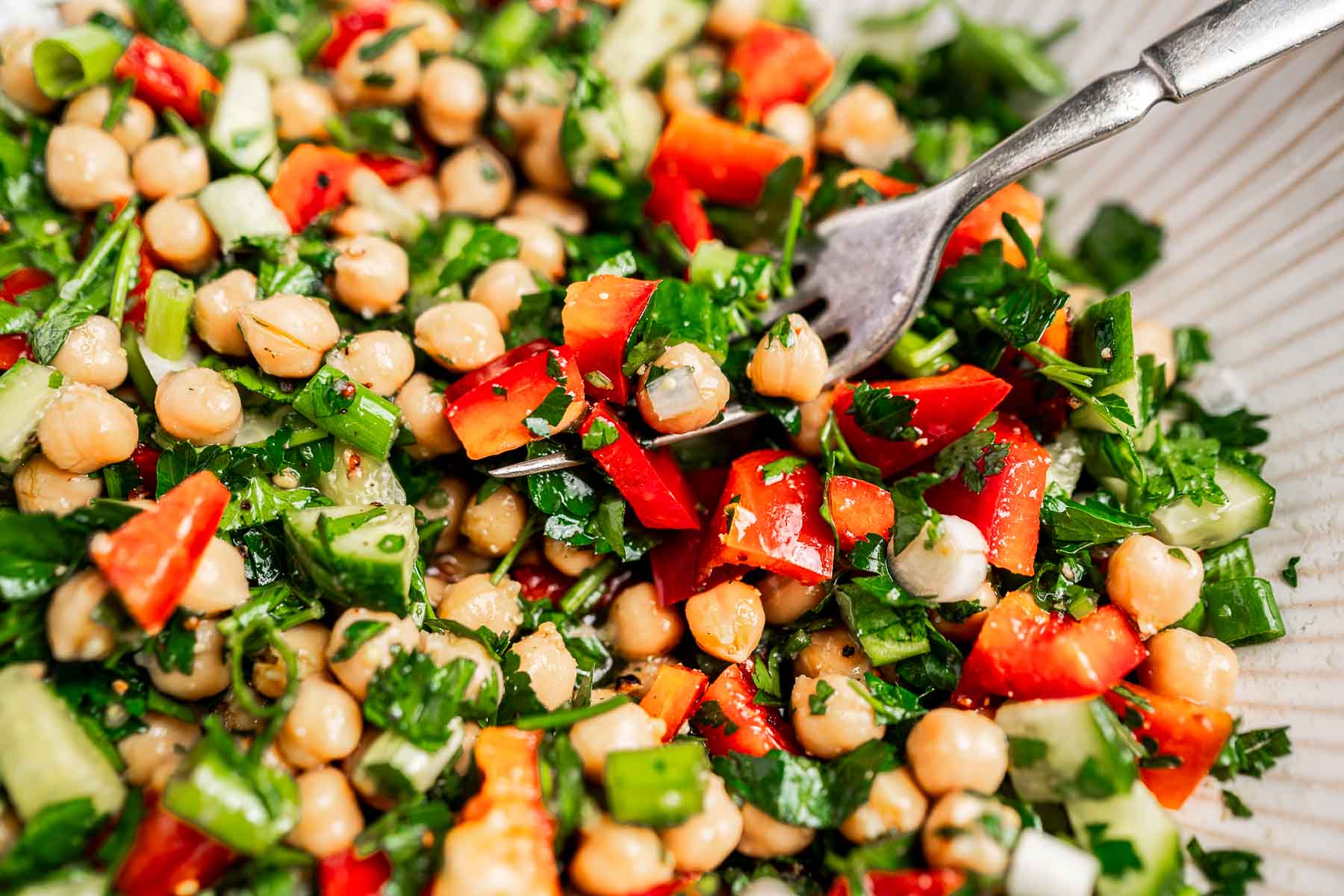 Close-up of a chopped chickpea salad featuring red bell peppers, cucumber, and parsley, with a fork on the side.