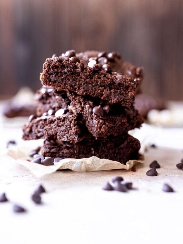 A stack of chocolate brownies with chocolate chips scattered around on a parchment paper.