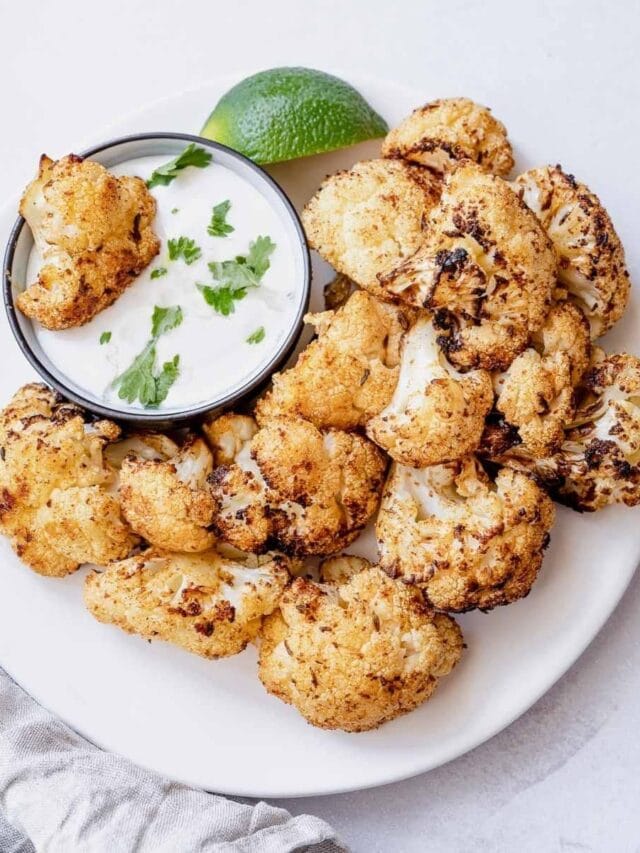 A plate of roasted cauliflower florets with a side of creamy dip garnished with cilantro, accompanied by a lime wedge.