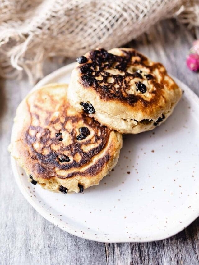 Two toasted crumpets with raisins on a white speckled plate, set on a wooden table with a burlap cloth in the background.