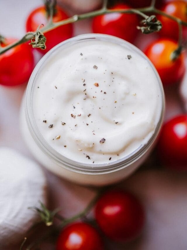 A jar of creamy white sauce sprinkled with black pepper, surrounded by red cherry tomatoes and garlic cloves.