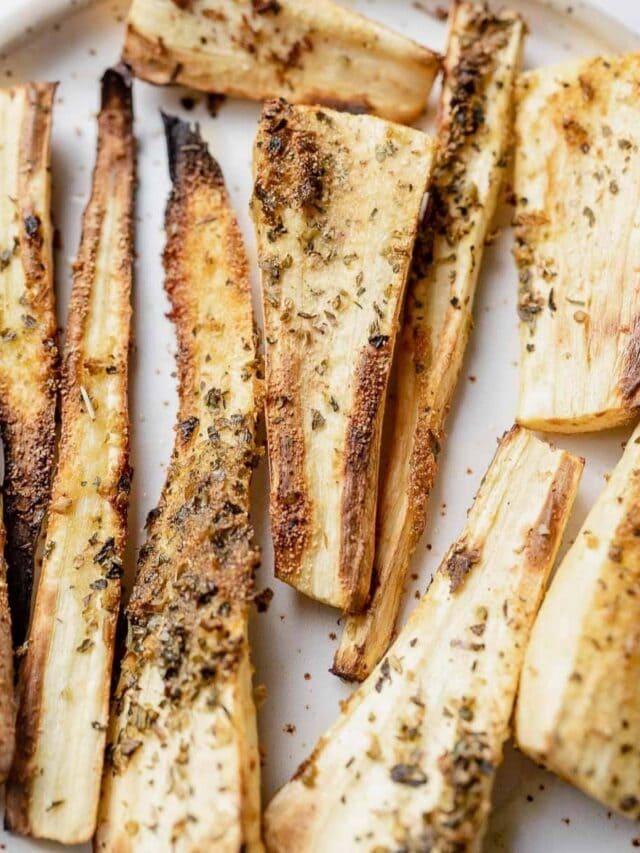 Close-up of roasted parsnip wedges on a white plate, seasoned with herbs and spices, showing a golden-brown, slightly charred exterior.