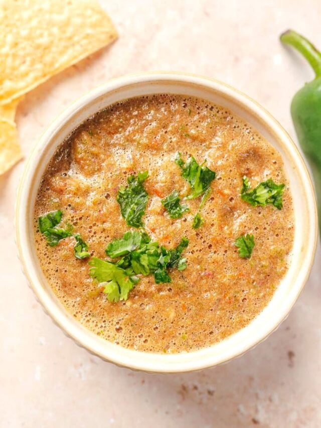 A bowl of salsa garnished with chopped cilantro. A tortilla chip and a jalapeño pepper are nearby.