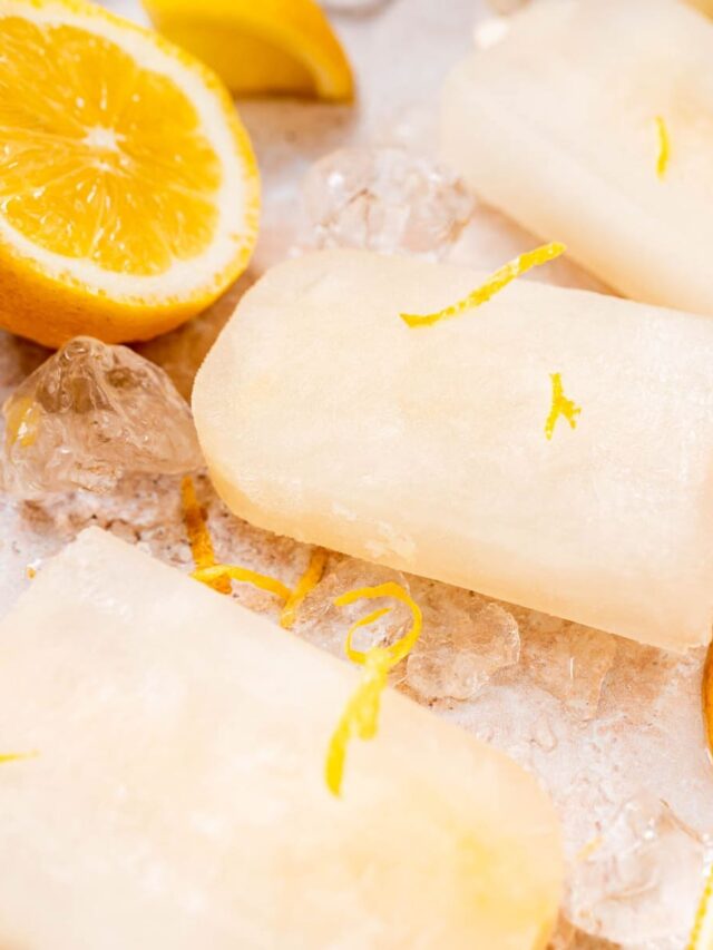 Lemon popsicles with zest and ice cubes on a surface, accompanied by halved lemons.