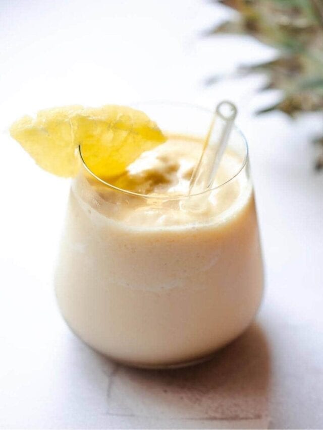 A creamy smoothie served in a glass with a clear straw, topped with a slice of pineapple, against a neutral background.