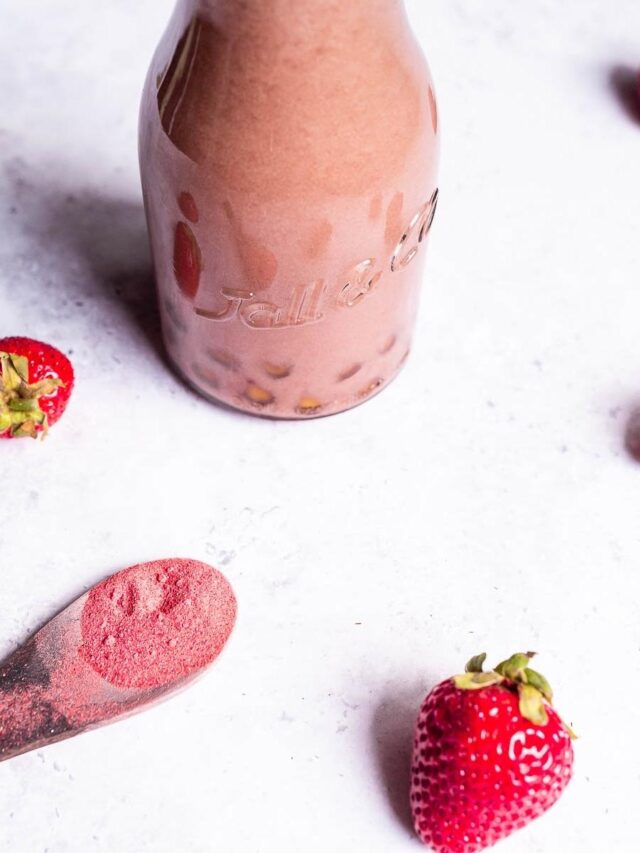 A bottle of chocolate milkshake on a white surface, surrounded by a whole strawberry, a wooden spoon with powder, and another strawberry partially visible.