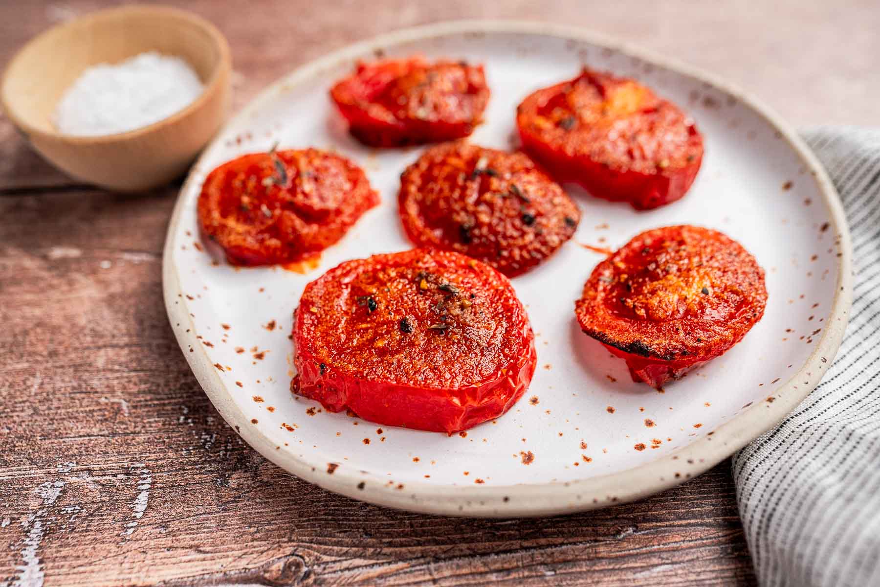 A plate of air fryer tomatoes sits beside a small bowl of salt on a wooden table topped with a striped cloth.