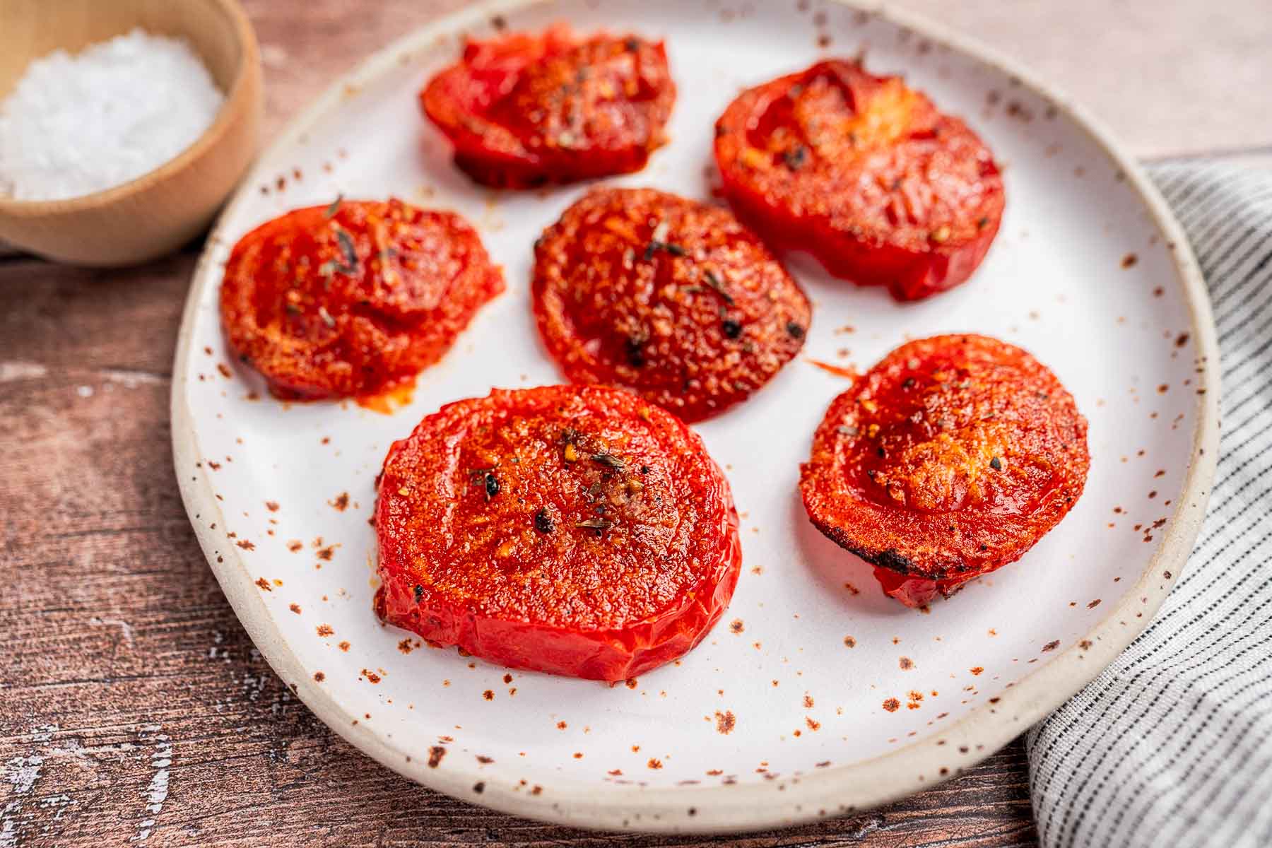Plate with six perfectly roasted air fryer tomatoes next to a small wooden bowl of salt on a wooden surface.