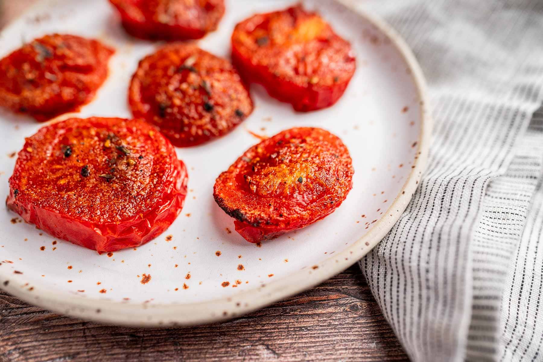 Slices of air fryer roasted tomatoes with seasoning rest on a white plate, beside a striped textile on a wooden surface.
