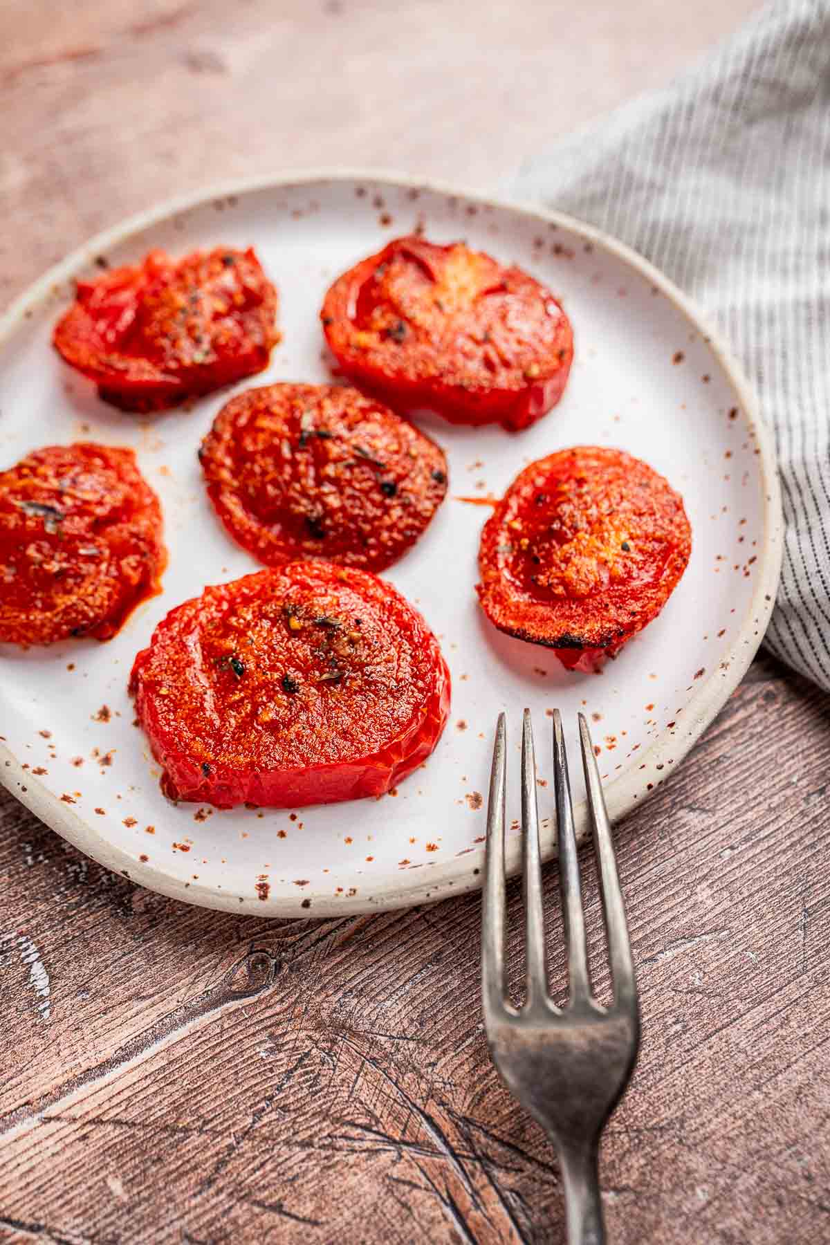 A plate of six air fryer tomatoes, perfectly seasoned and grilled, sits on a white dish with a metal fork beside it on a wooden surface.