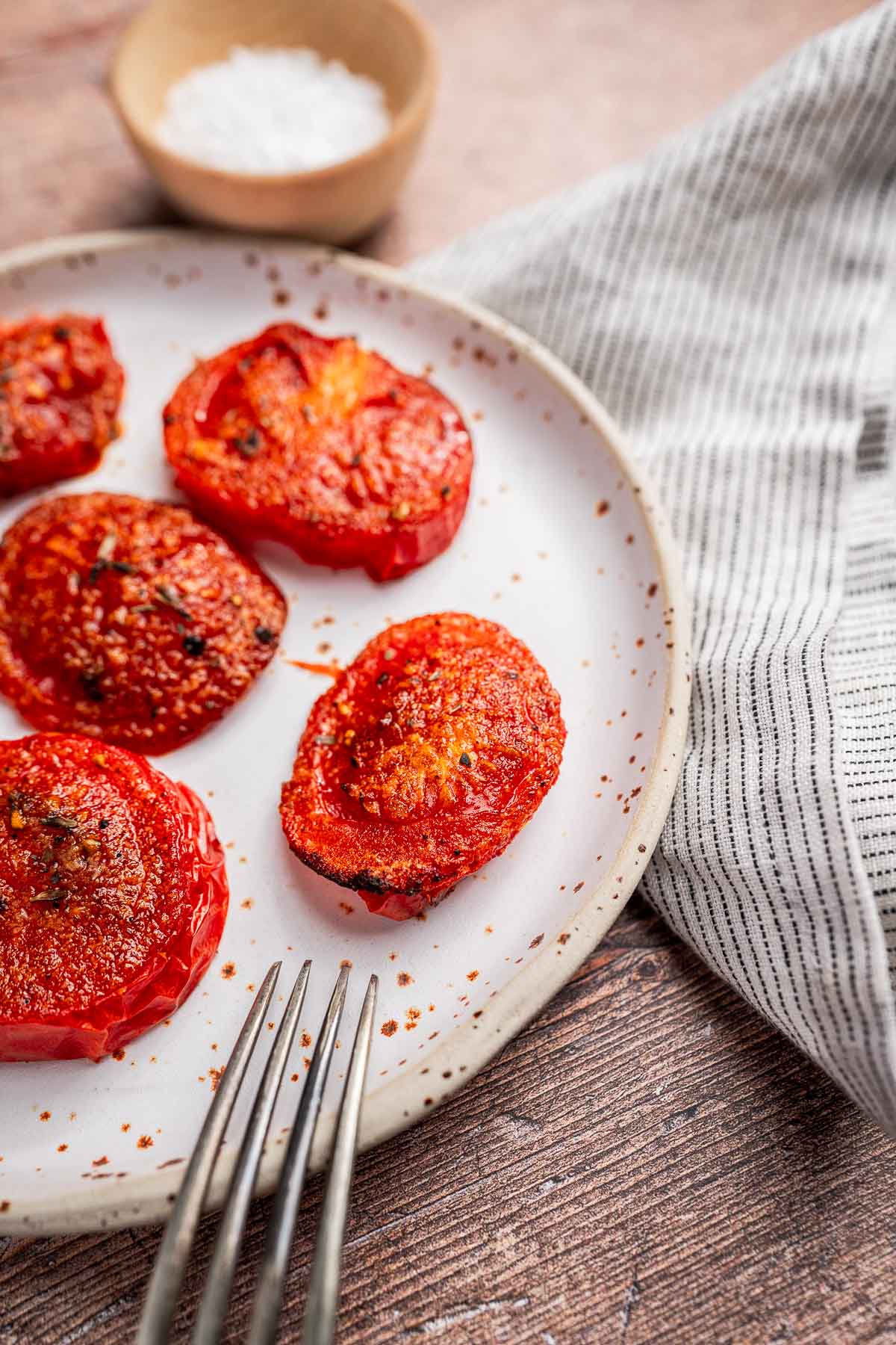 A plate of air fryer tomatoes seasoned with spices, a fork nearby, a cloth napkin, and a small bowl of salt in the background.