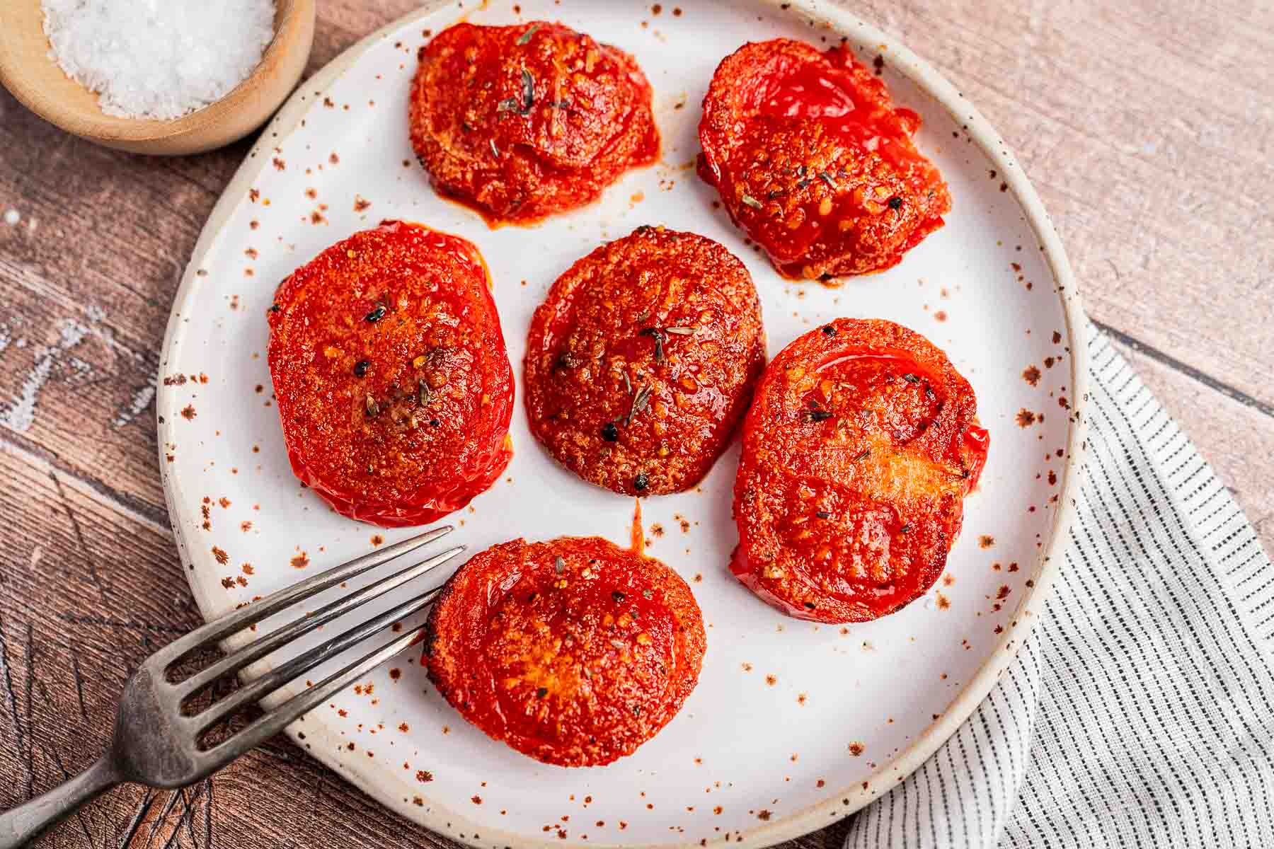 Plate with six roasted air fryer tomato slices seasoned with herbs, a fork on the side, and a striped napkin nearby.