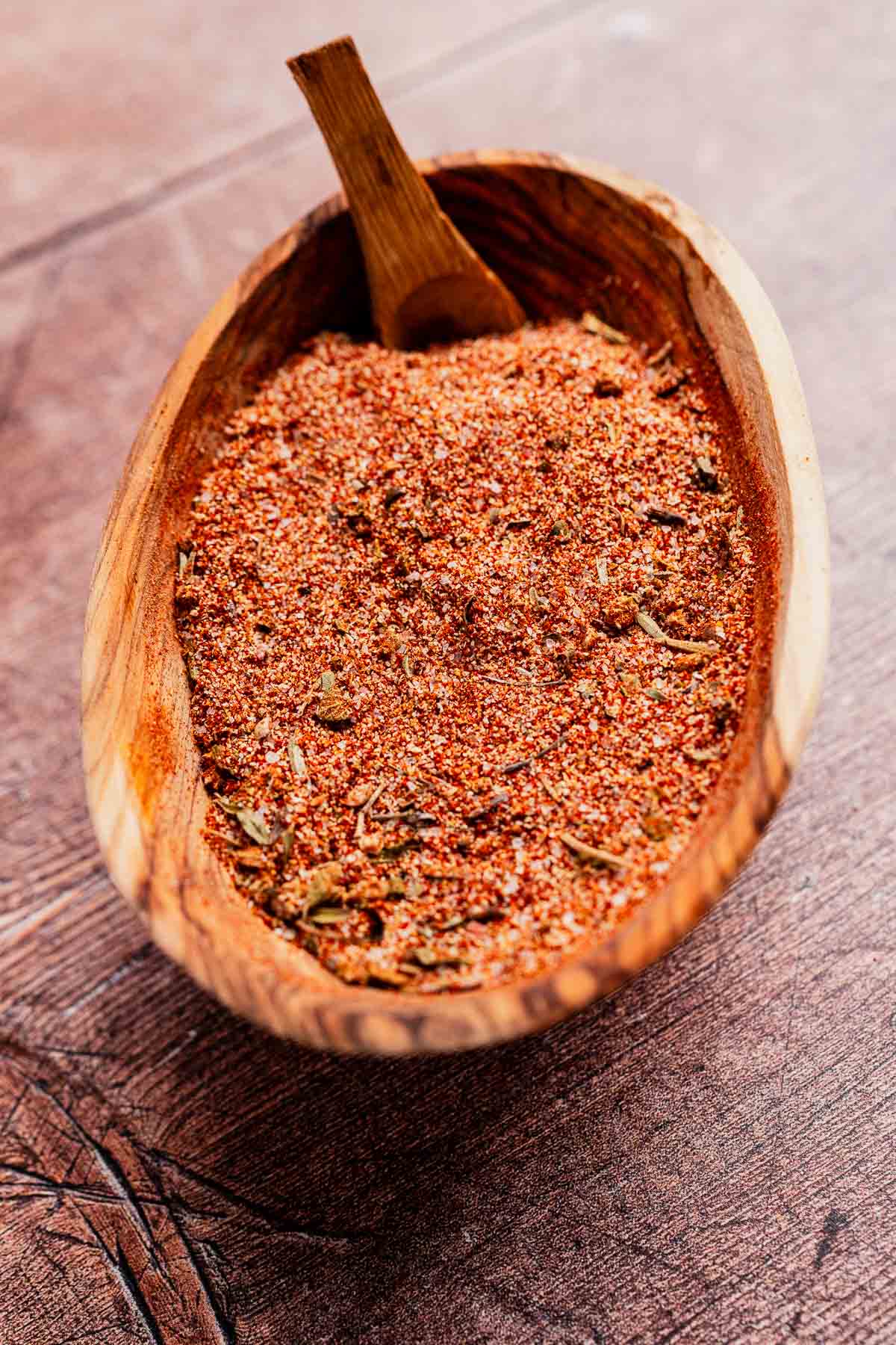 A wooden bowl brimming with Cajun seasoning and an assortment of herbs, accompanied by a small wooden scoop, rests elegantly on a wooden surface.