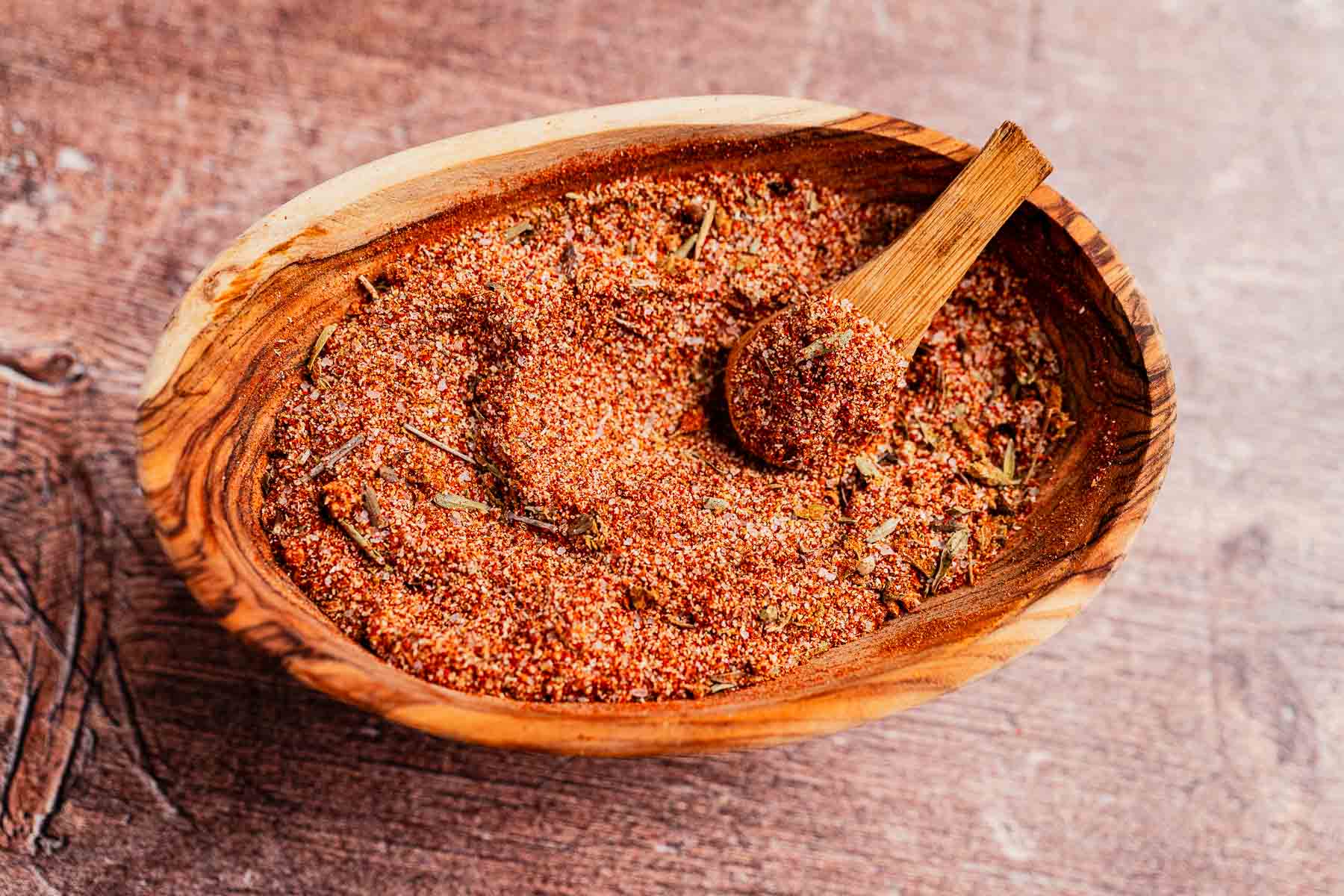 A wooden bowl filled with a vibrant Cajun seasoning mix, featuring an array of red spices and herbs, complete with a small wooden scoop.