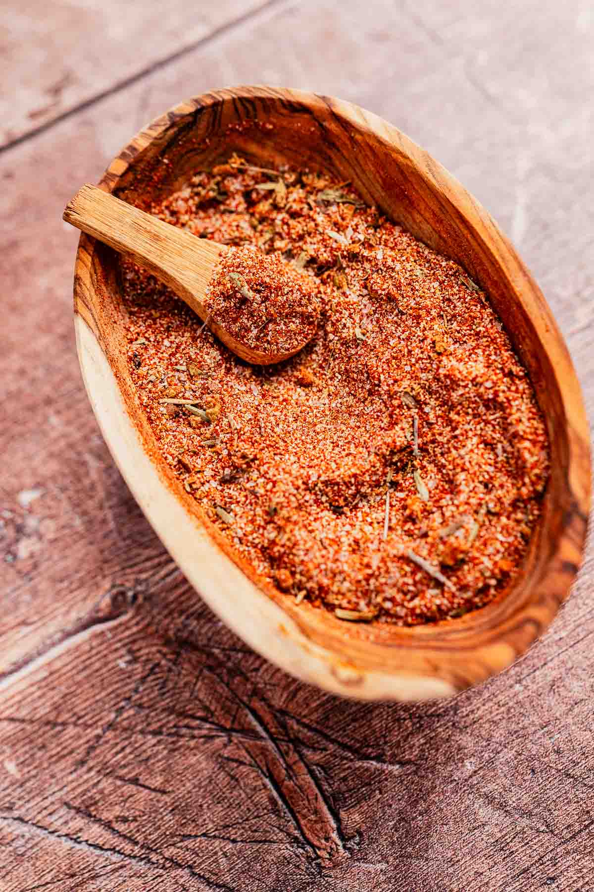 A wooden bowl brimming with cajun seasoning and a small wooden spoon rests on a textured wooden surface.