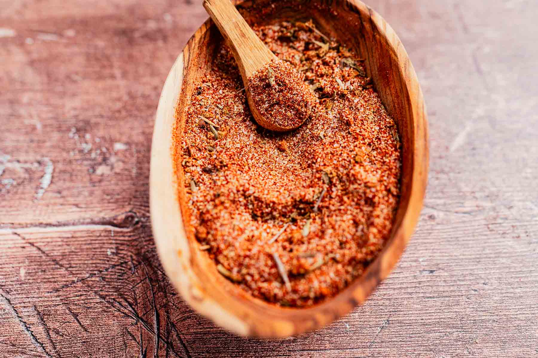 A wooden bowl filled with a vibrant Cajun seasoning mix and a small wooden spoon rests on a rustic wooden surface.