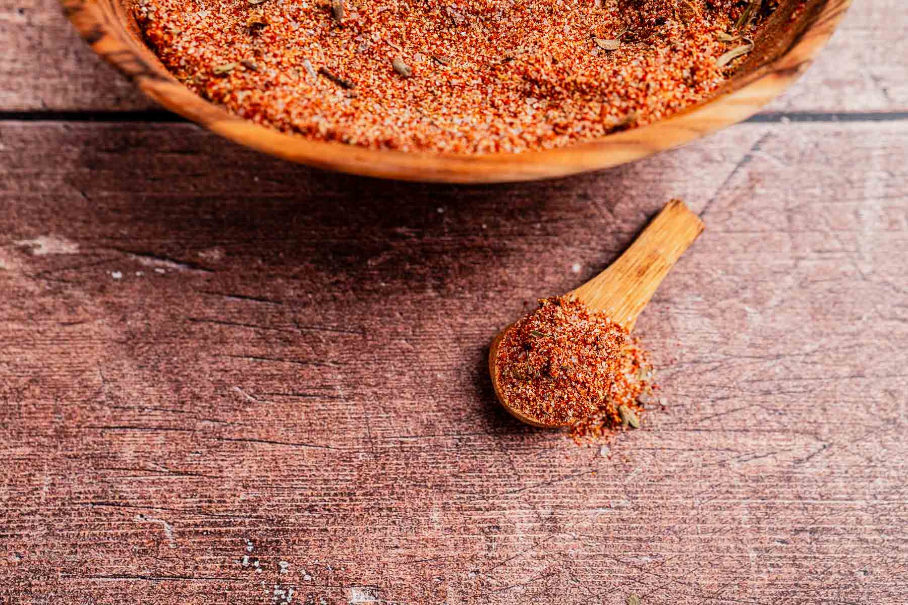 A wooden bowl and spoon filled with a rich, reddish-brown Cajun seasoning mix sit on a wooden table.