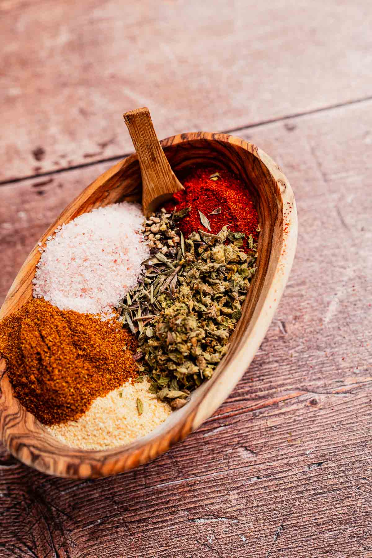 A wooden bowl with sections of various spices: paprika, salt, herbs, curry powder, cajun seasoning, and other flavorful seasonings sits on a wooden table.