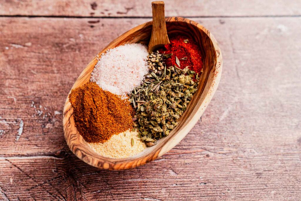 A wooden bowl holds assorted spices: red chili powder, dried herbs, coarse salt, and curry powder, with a hint of Cajun seasoning all placed on a rustic wooden surface.