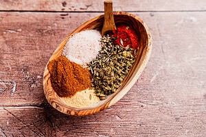 A wooden bowl holds assorted spices: red chili powder, dried herbs, coarse salt, and curry powder, with a hint of Cajun seasoning all placed on a rustic wooden surface.