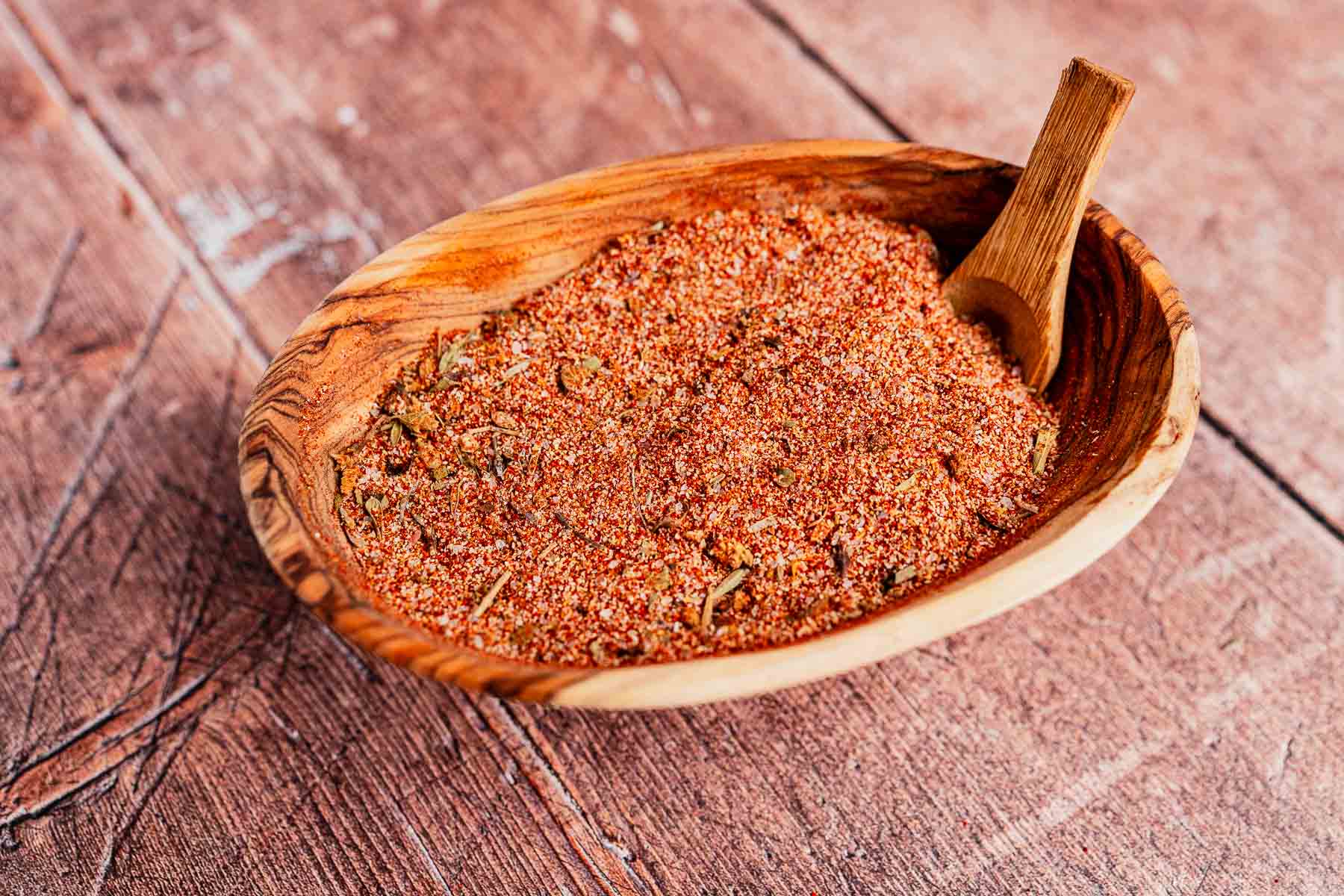 A wooden bowl filled with a vibrant Cajun seasoning mix, accompanied by a small wooden spoon, rests elegantly on a rustic wooden surface.