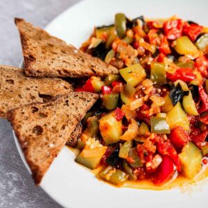 A plate of ratatouille with diced zucchini, bell peppers, onions, and tomatoes, accompanied by three triangular slices of whole grain bread.