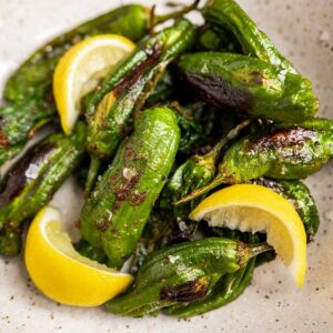 A plate of grilled shishito peppers with char marks, garnished with lemon wedges.