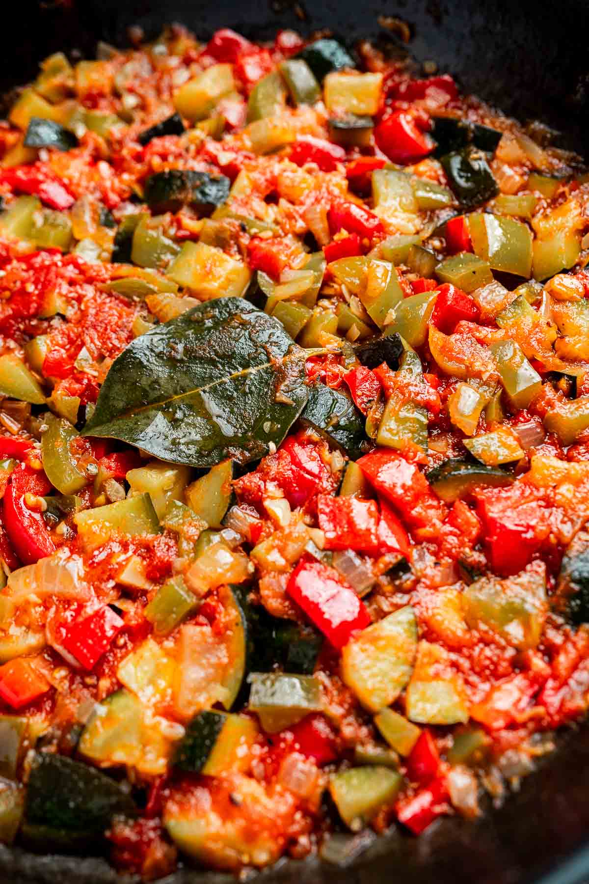Close-up of a colorful pisto, a vegetable stew with chopped tomatoes, zucchini, bell peppers, and a bay leaf, all simmering together in a pan.