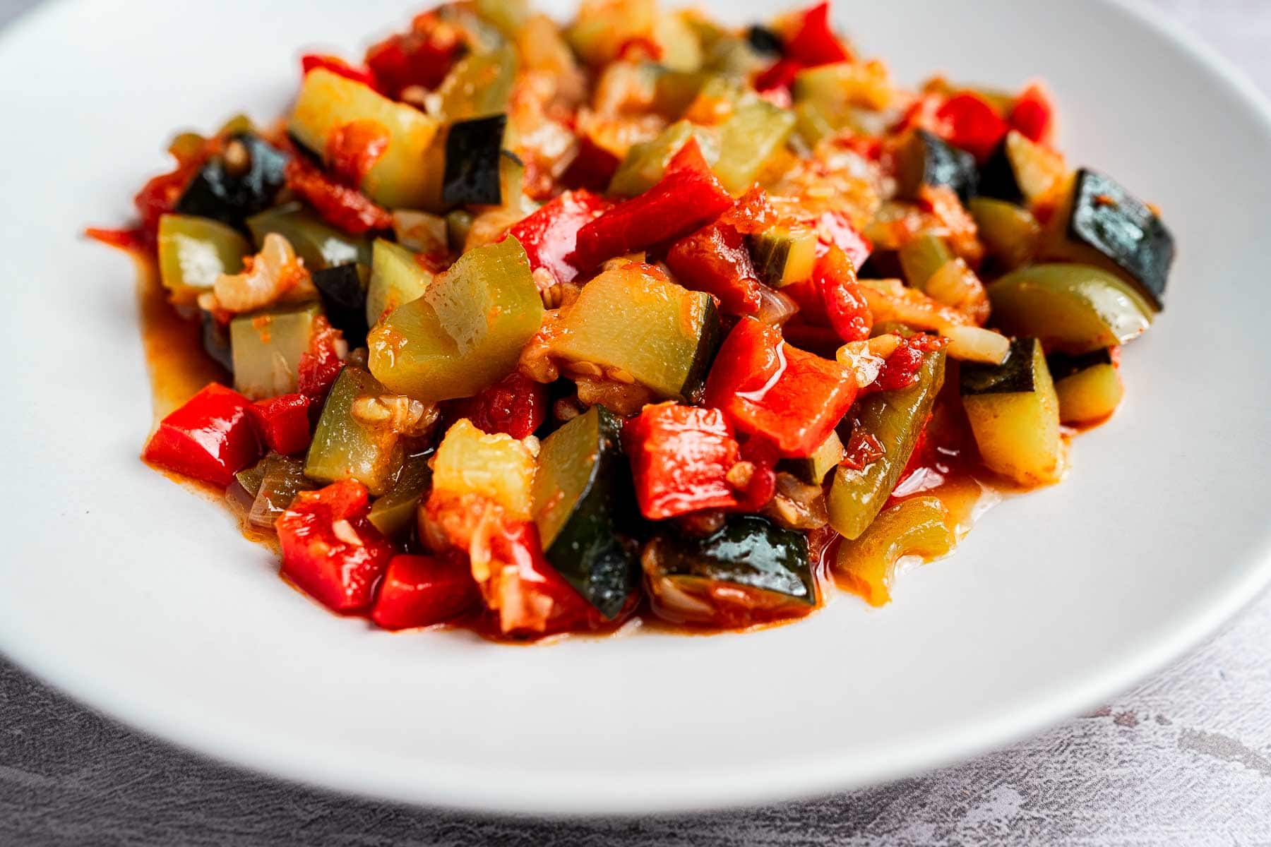 A white plate filled with a colorful vegetable dish featuring red and green peppers, zucchini, and other mixed vegetables in a sauce.