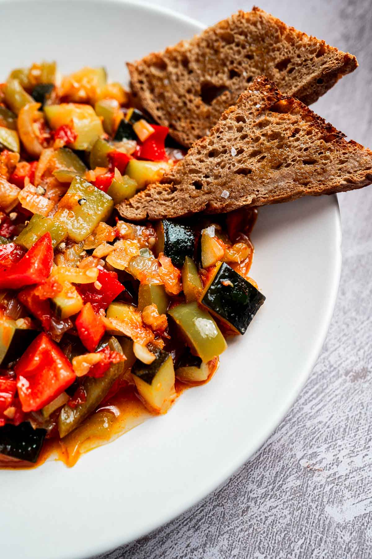 A white plate features a vibrant pisto with a vegetable medley of zucchini, red and green bell peppers, and onions, accompanied by two slices of brown bread.