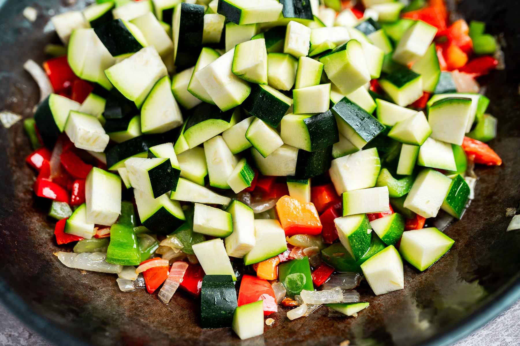Chopped zucchini, red peppers, and onions are sautéing in a dark skillet.
