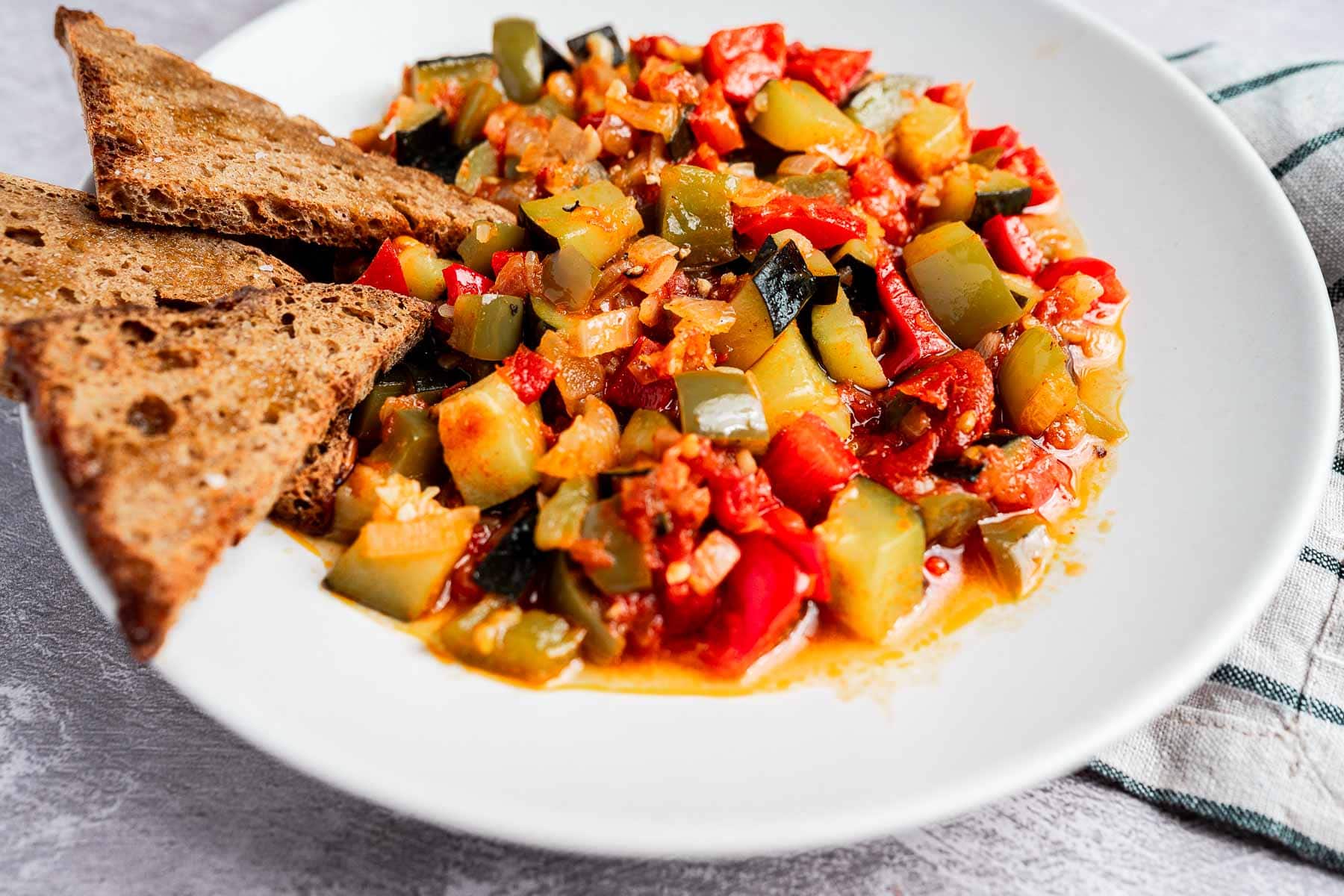 A white plate with ratatouille, reminiscent of pisto, features vibrant chopped vegetables in a rich tomato-based sauce, accompanied by two pieces of toasted brown bread.