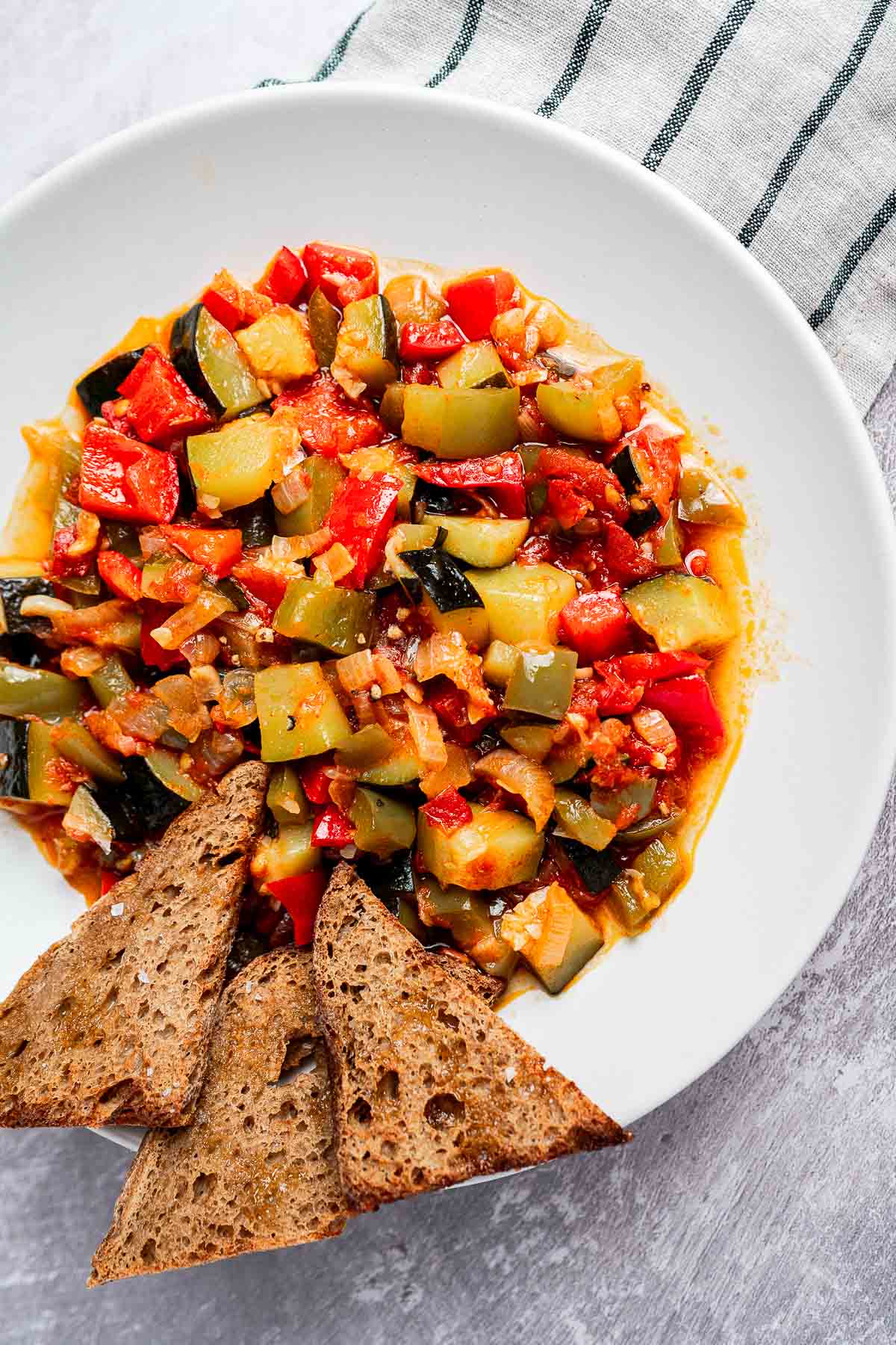 A plate of colorful ratatouille, reminiscent of traditional pisto, with diced vegetables on a white plate, accompanied by three triangular slices of brown bread.