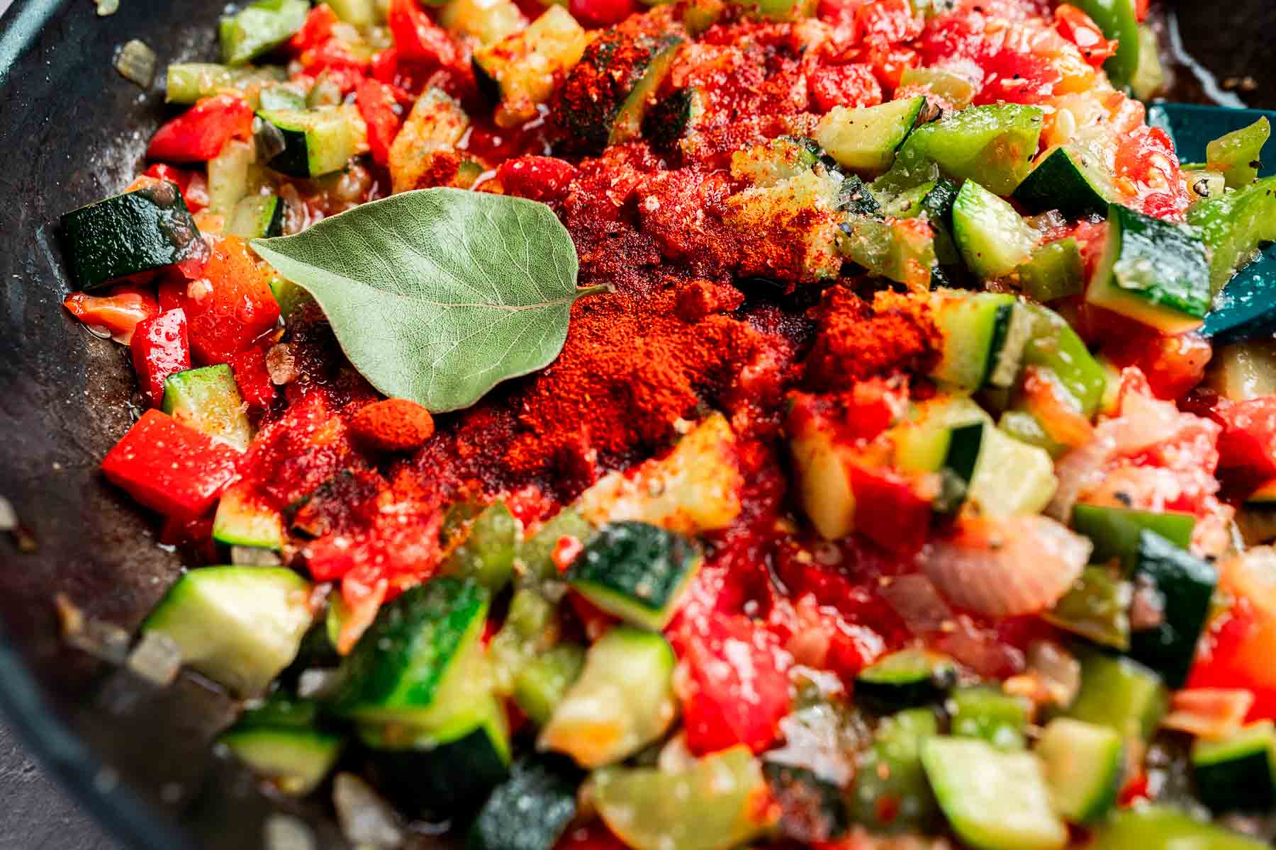 Close-up of a colorful vegetable dish in a pan, featuring chopped zucchini, red bell peppers, onion, a bay leaf, and a sprinkle of red spices.