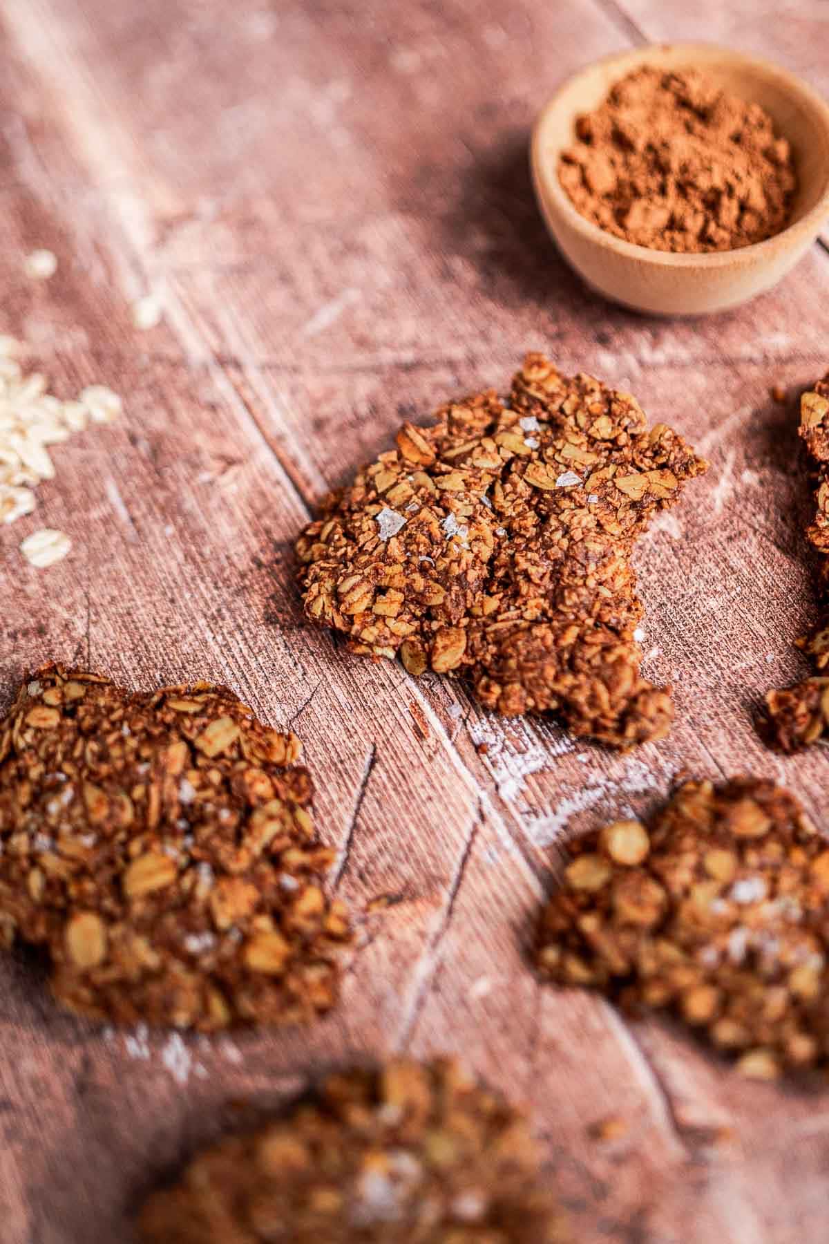Close-up of broken oat cookies on a wooden surface with a small bowl of cocoa powder nearby.