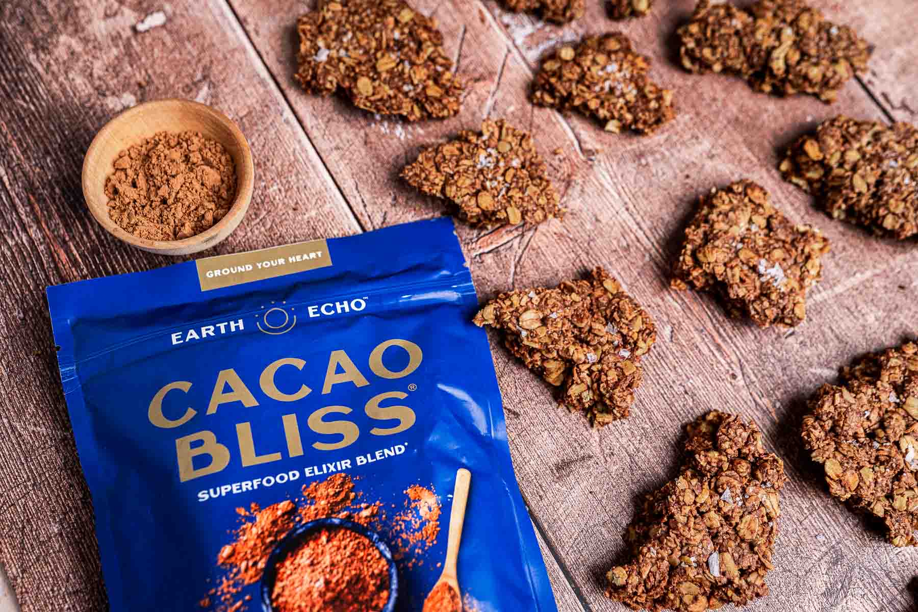 Blue Cacao Bliss packaging next to clusters of oat-filled snack pieces on a wooden surface, with a small bowl of loose blend.