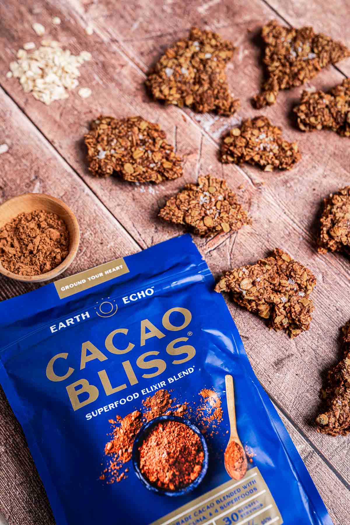 A blue Cacao Bliss package on a wooden surface, surrounded by broken pieces of baked chocolate granola and a small wooden bowl of cocoa powder.