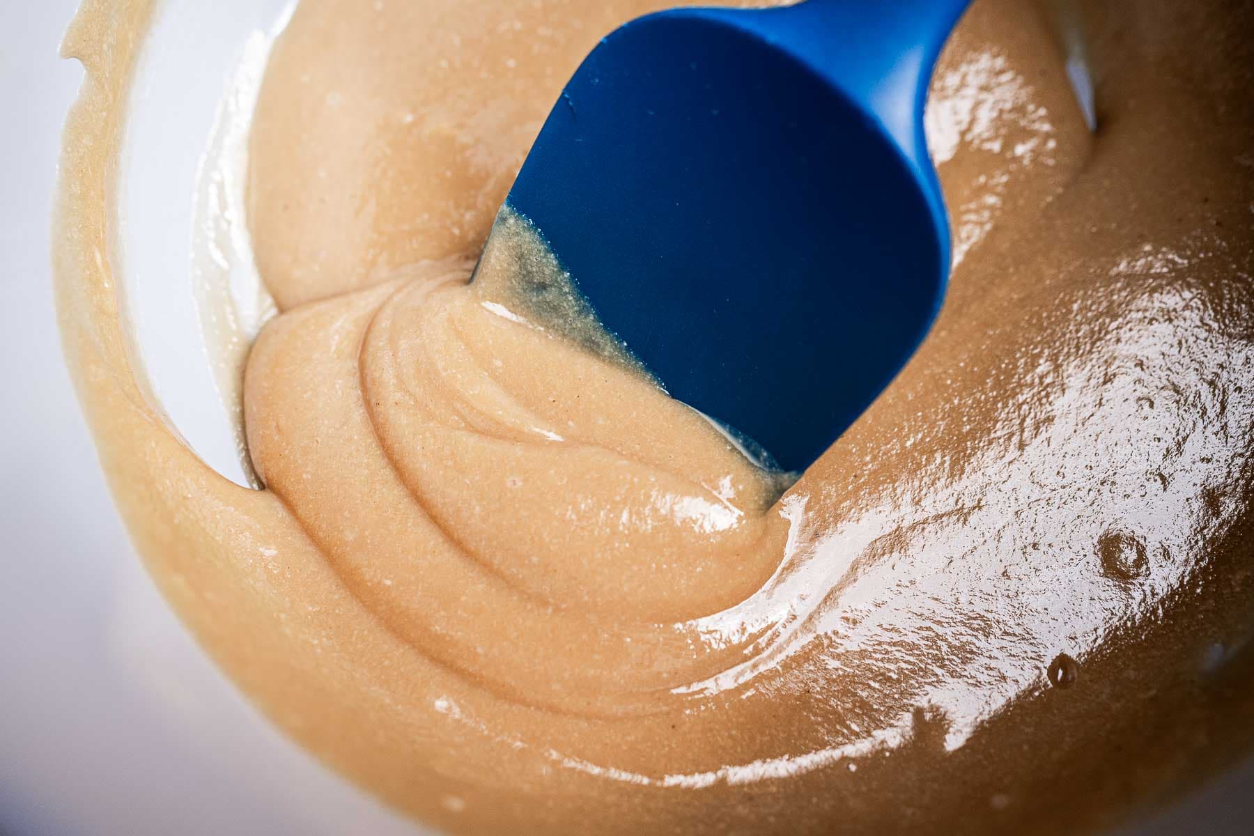 A blue spatula stirring smooth, creamy peanut butter in a bowl.