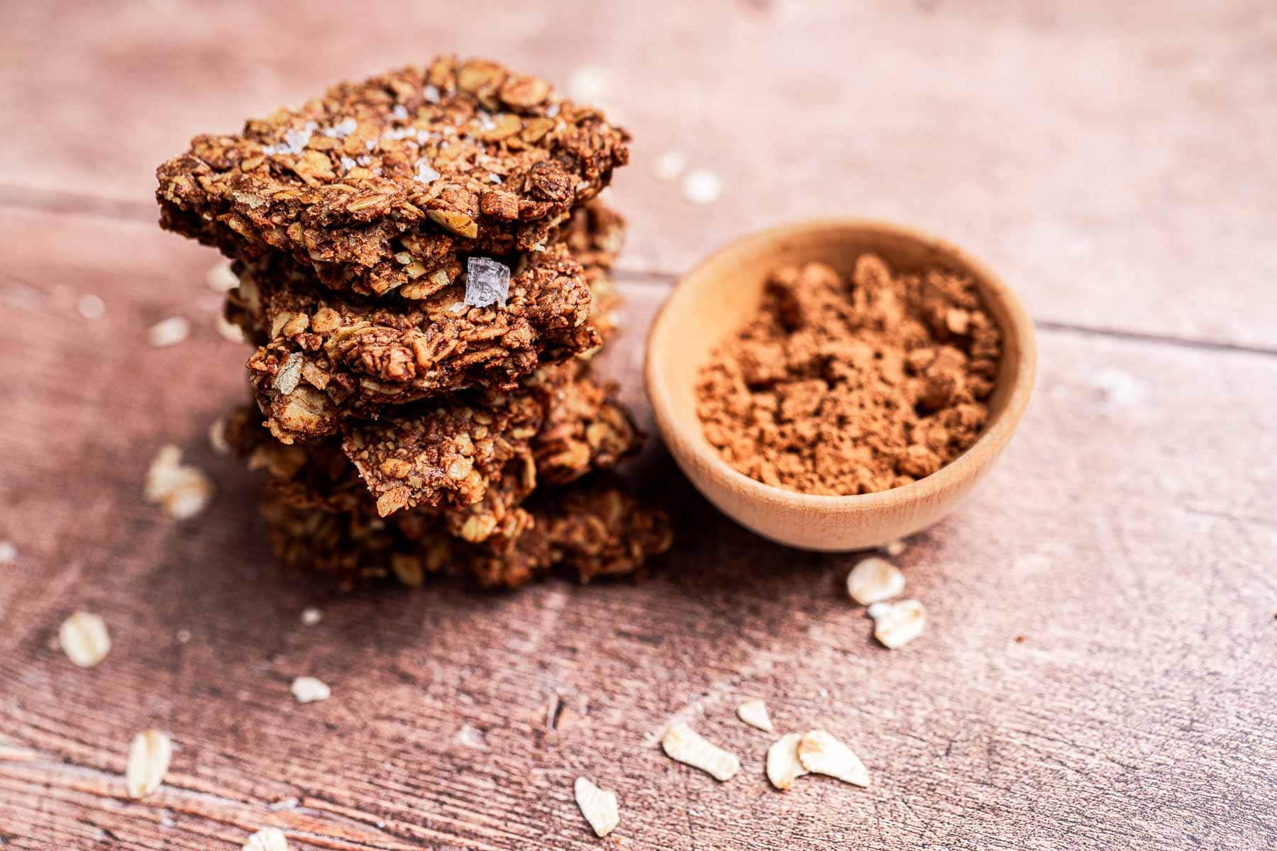 Stack of homemade oat cookies with a sprinkle of salt next to a wooden bowl containing brown sugar, on a wooden surface.
