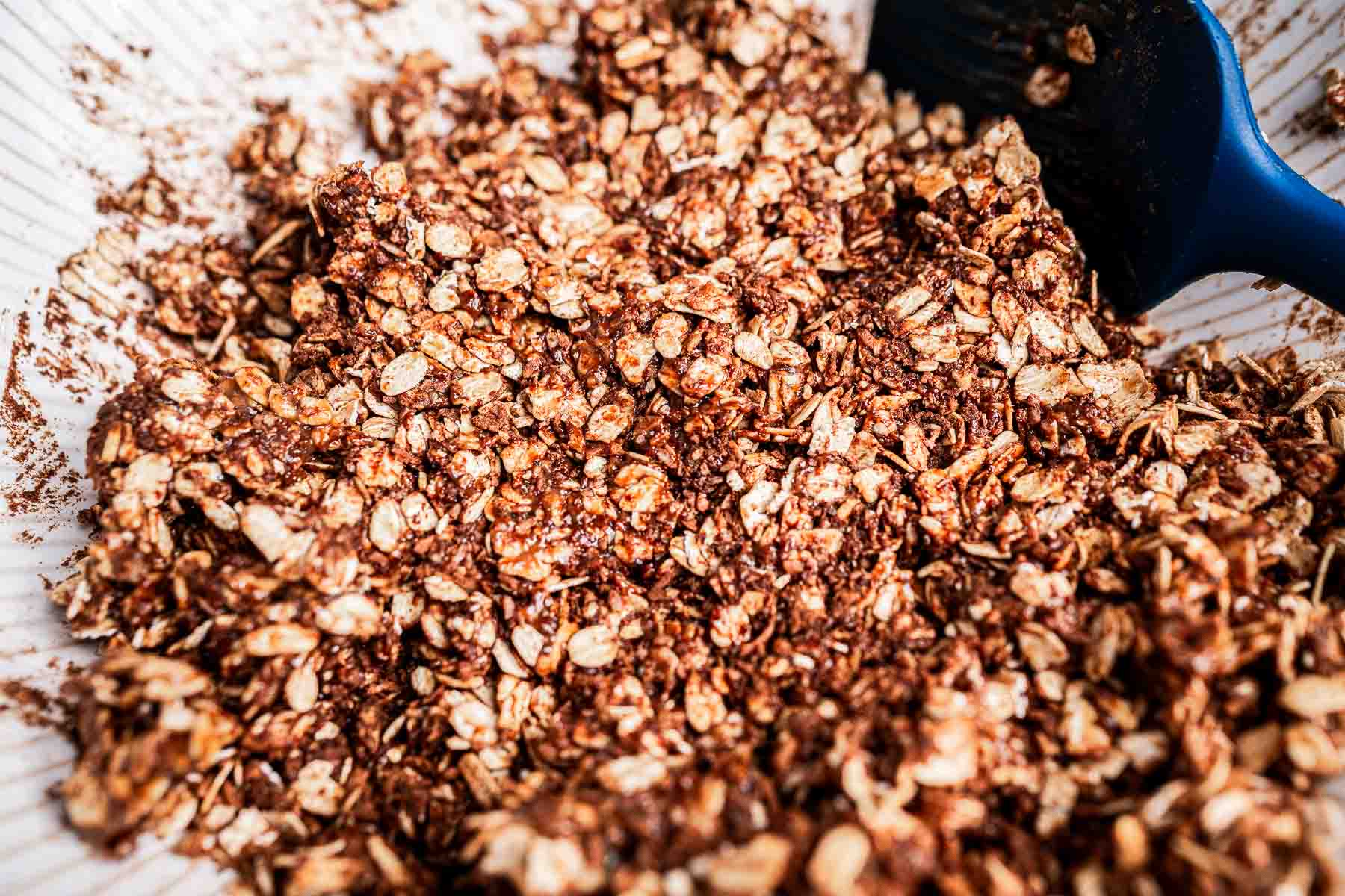 A close-up of granola mixed with cocoa powder in a bowl, with a blue spatula partially in view on the right side.
