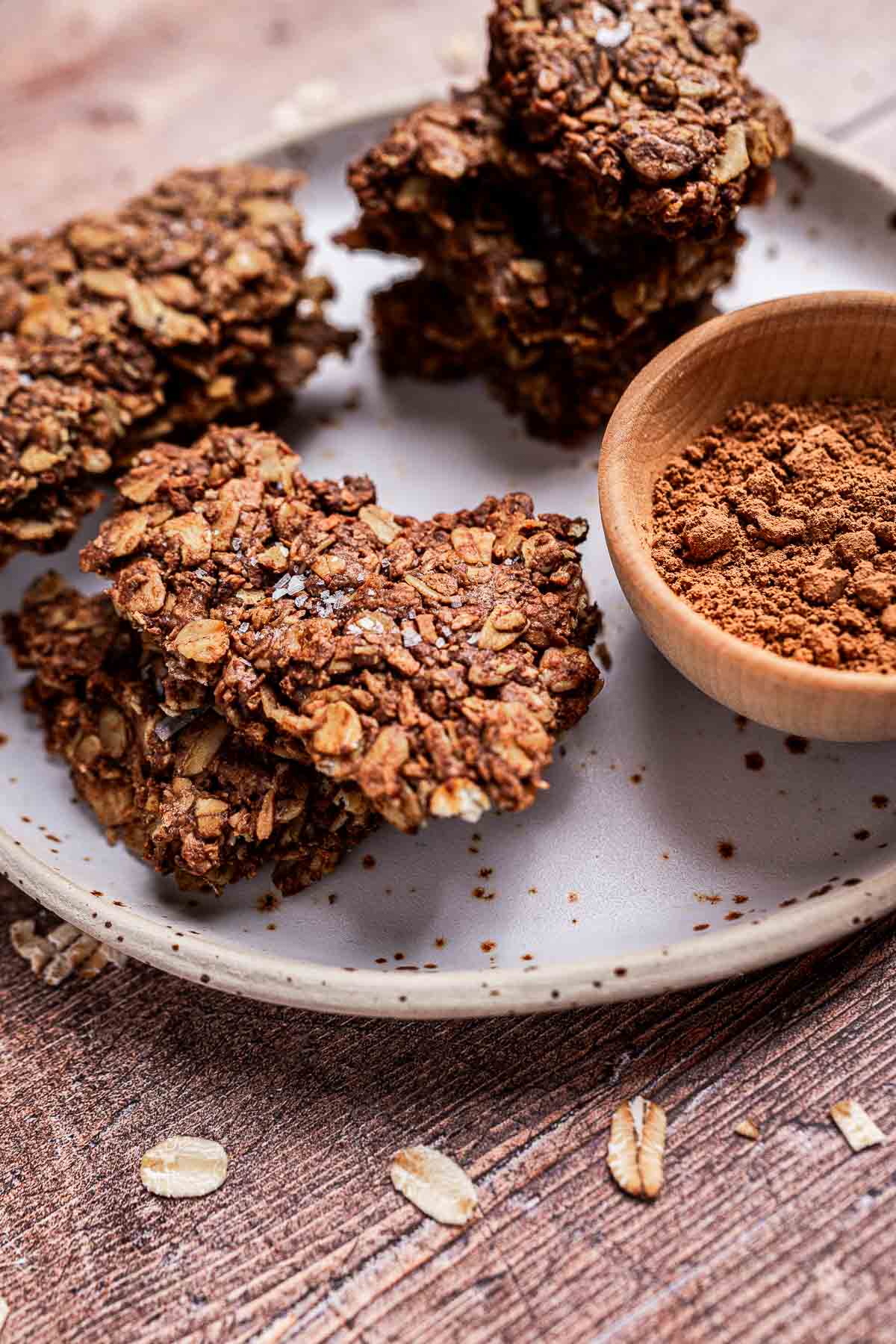 A plate with oat and chocolate chip cookies next to a wooden bowl of cocoa powder.