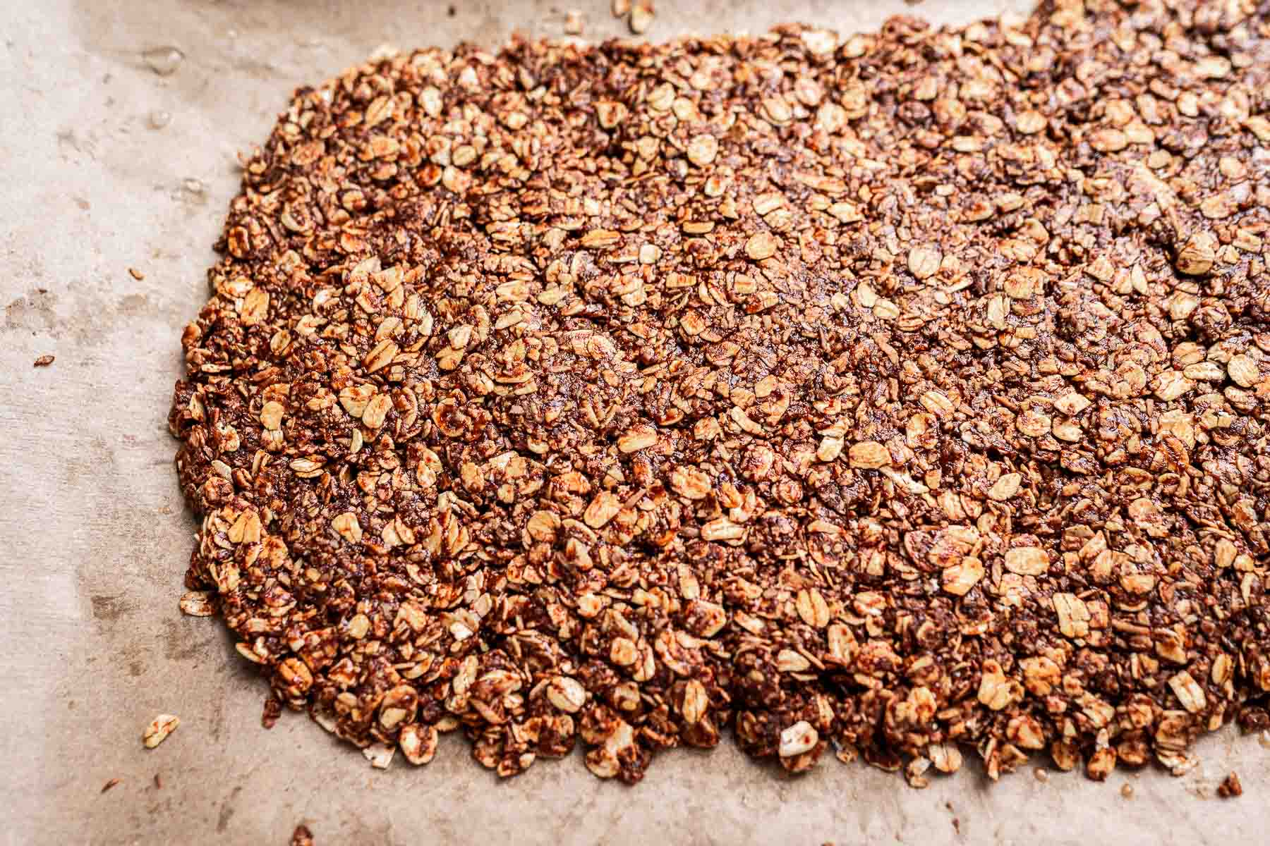 A baked sheet of homemade chocolate granola spread out unevenly on parchment paper.