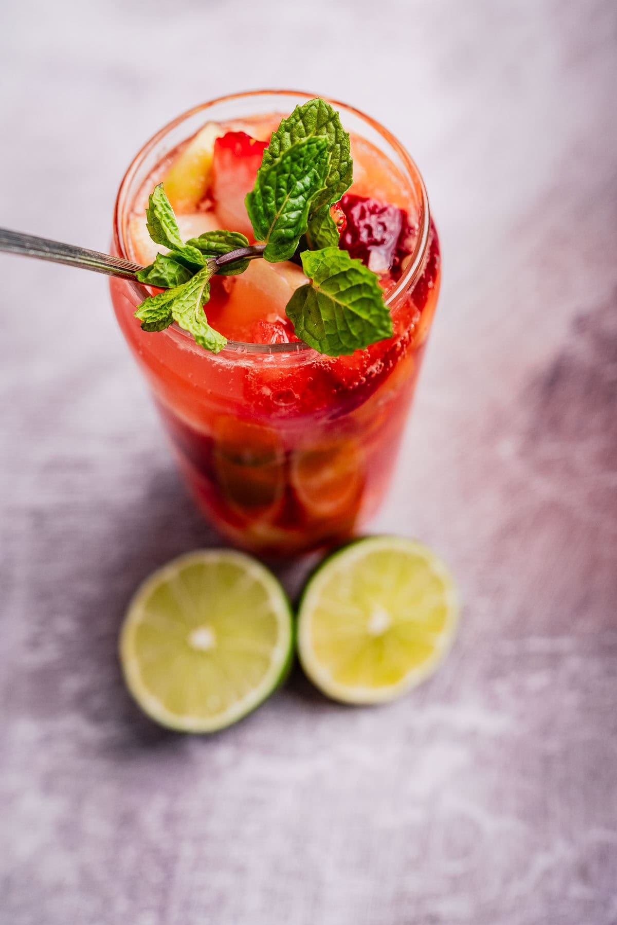 A refreshing tizana in a glass with strawberries, mint, and ice. Two halved limes are placed beside the glass on a gray surface.