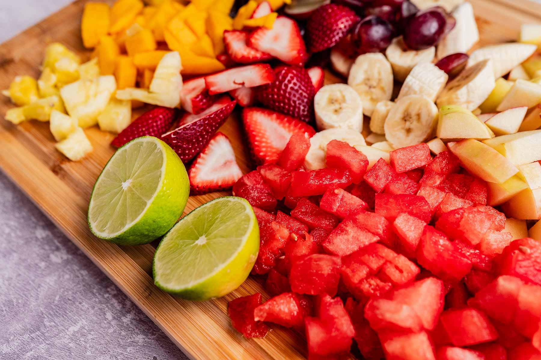 A wooden board with an assortment like a tizana: pineapple, strawberries, bananas, apples, grapes, watermelon, and two lime halves.